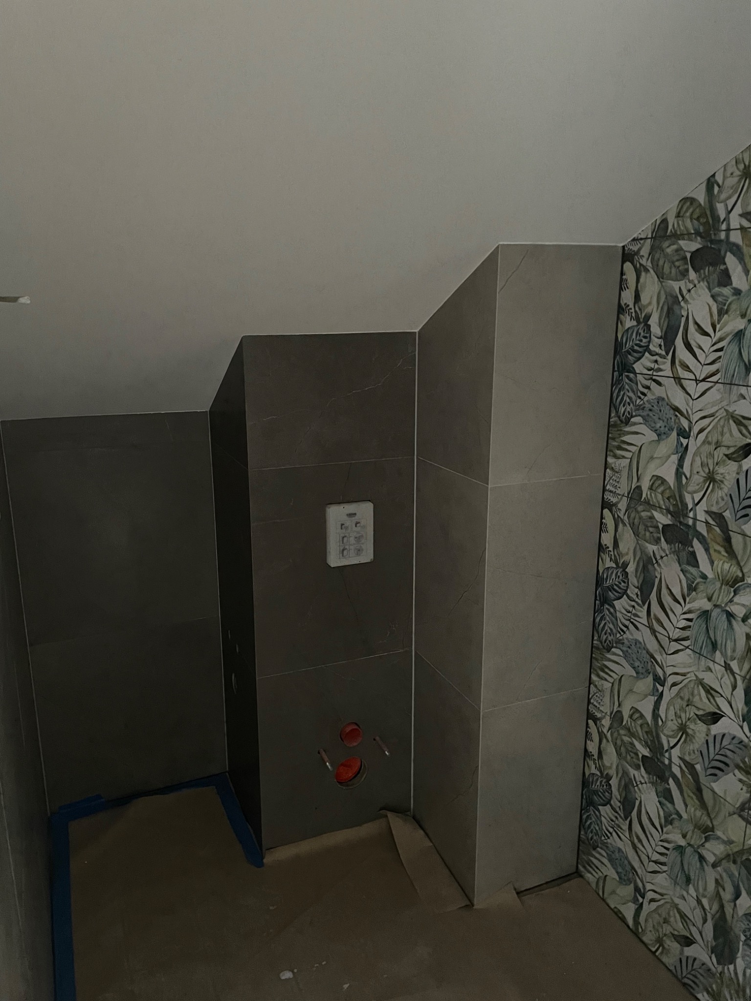 Wnętrze łazienki w trakcie remontu: szare płytki imitujące marmur na ścianach, fragment ściany z tapetą w motywy roślinne, wystające rury, zabezpieczona podłoga.