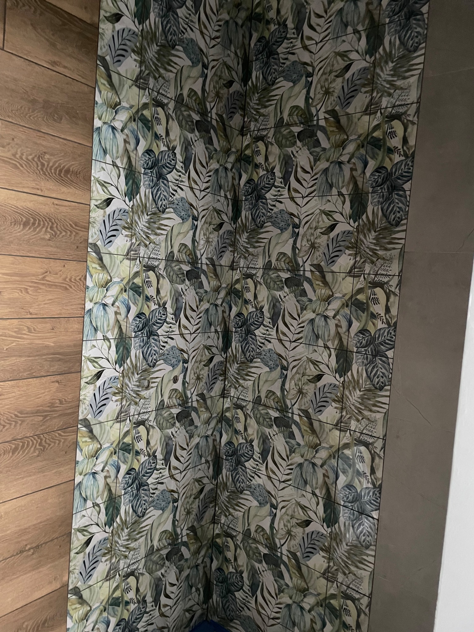 Ściana łazienki wykończona płytkami z motywem roślinnym, sąsiadująca z drewnopodobną okładziną i szarą ścianą.