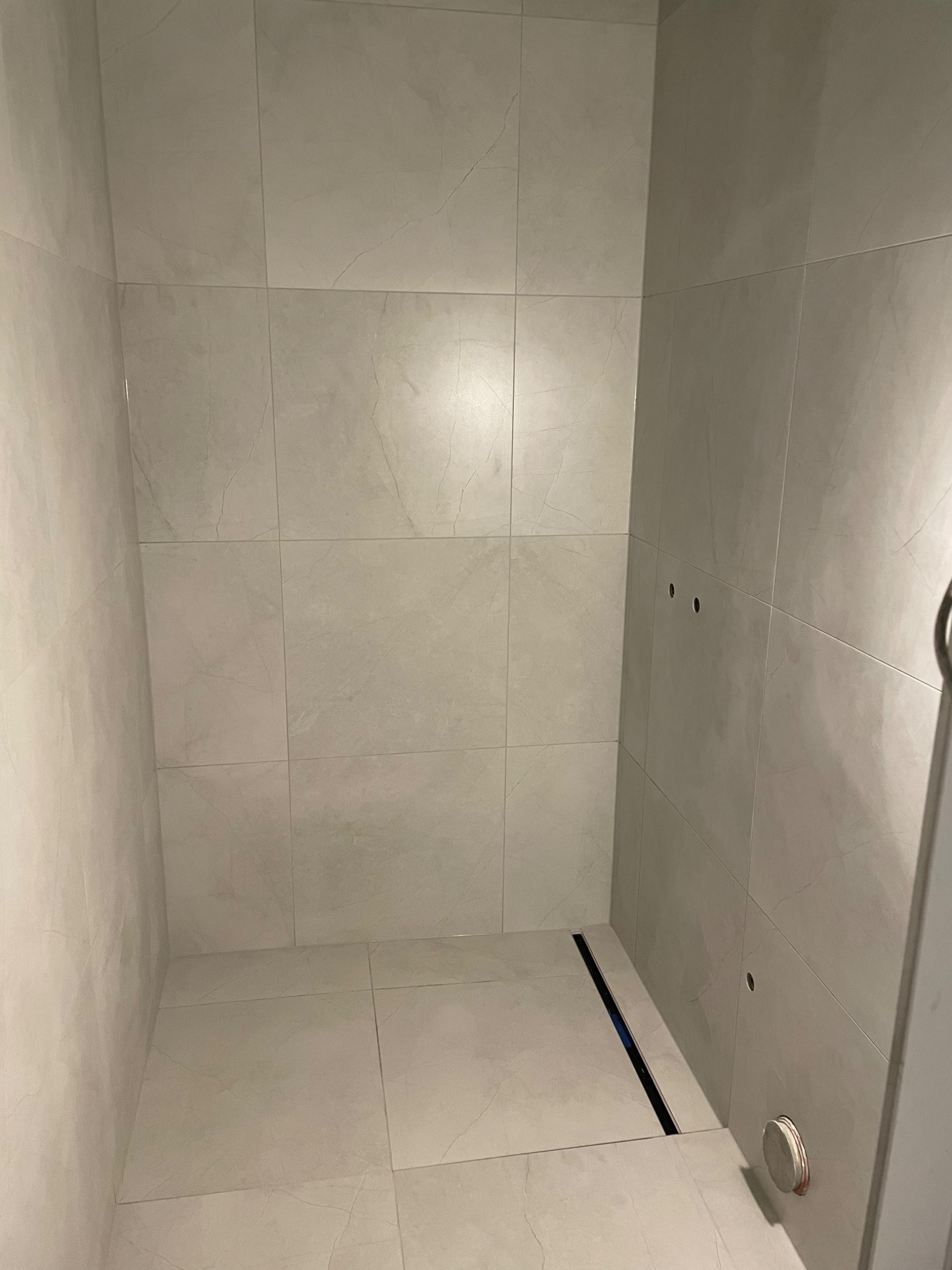 Wykończony narożnik kabiny prysznicowej z jasnymi, marmurkowymi płytkami ceramicznymi na ścianach i podłodze, widoczny odpływ liniowy oraz otwory na armaturę.