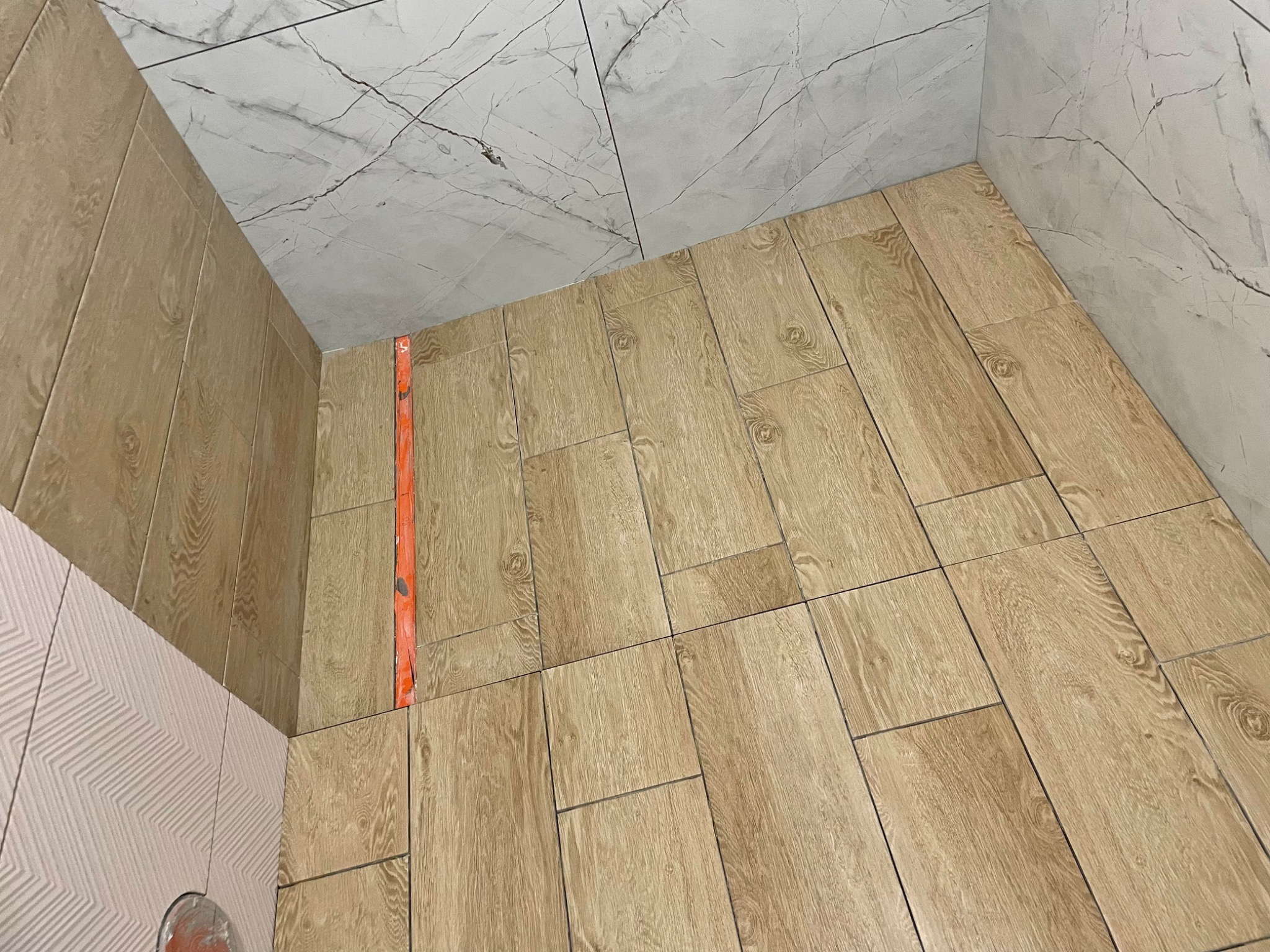 Układanie podłogowych płytek imitujących drewno w łazience, z poziomnicą przy ścianie i fragmentem ściany z białymi płytkami imitującymi marmur oraz różowymi płytkami z geometrycznym wzorem.