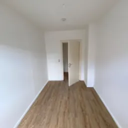 Wnętrze pokoju po remoncie z położonymi panelami podłogowymi o wzorze drewna i świeżo pomalowanymi na biało ścianami. Widoczny otwarty fragment drzwi.