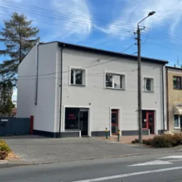 Dwukondygnacyjny budynek po odnowieniu elewacji, pomalowany na szaro z białymi obramowaniami okien i czerwonymi drzwiami wejściowymi, widoczny z poziomu ulicy w słoneczny dzień.