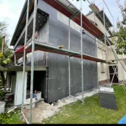 Dom w trakcie ocieplania: ściany pokryte czarnym styropianem, rusztowanie na dwóch ścianach, widoczne wiaderka z klejem i resztki materiałów budowlanych na trawie.