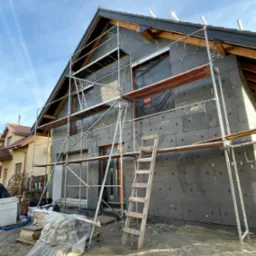 Dom jednorodzinny w trakcie ocieplania, widoczne rusztowanie, czarne płyty izolacyjne na ścianach, okna zabezpieczone folią, drewniana drabina oparta o rusztowanie, materiały budowlane na palecie...