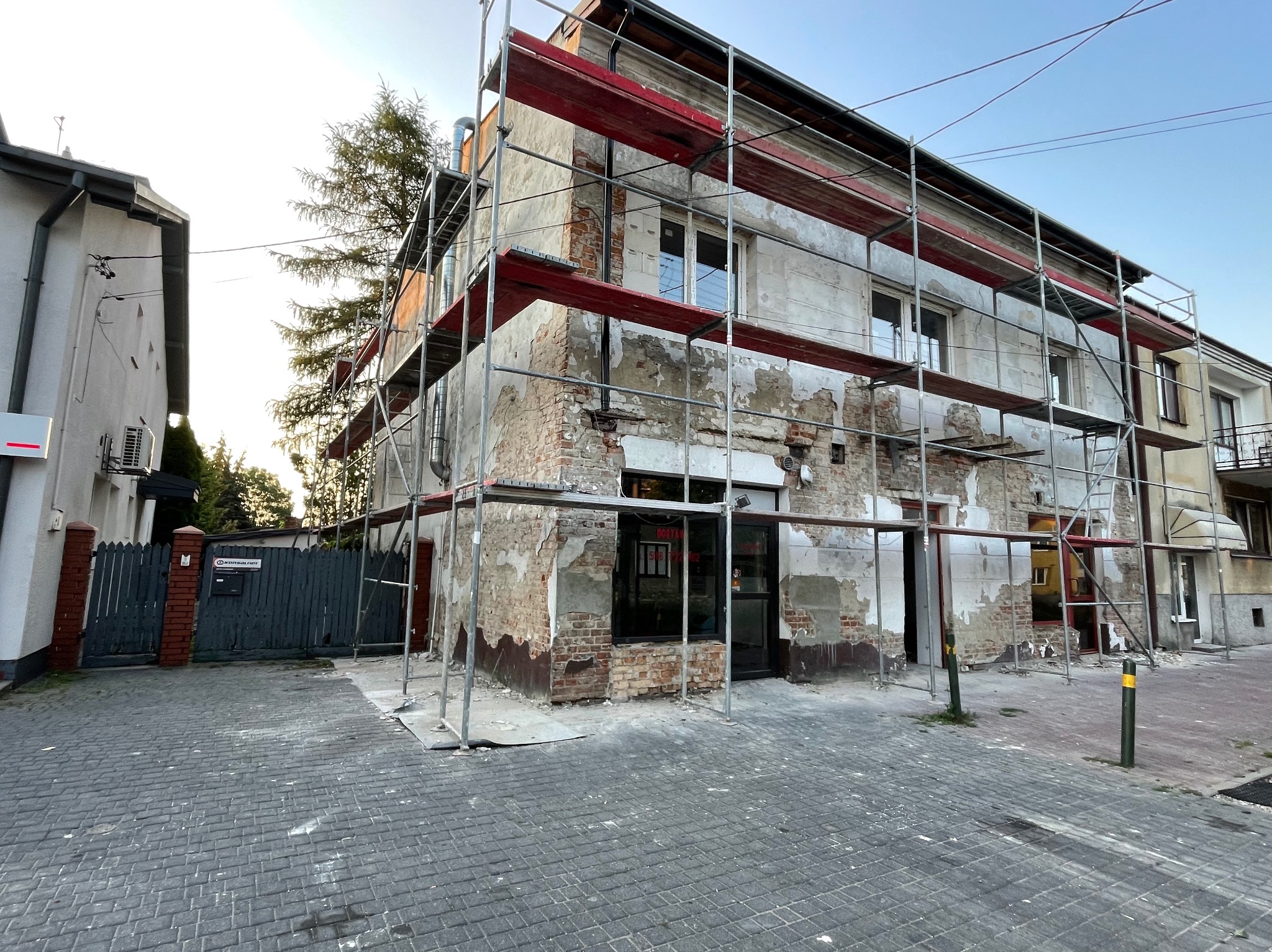 Budynek w Grądach w trakcie renowacji elewacji, widoczne rusztowania i fragmenty odsłoniętej cegły pod starym tynkiem.