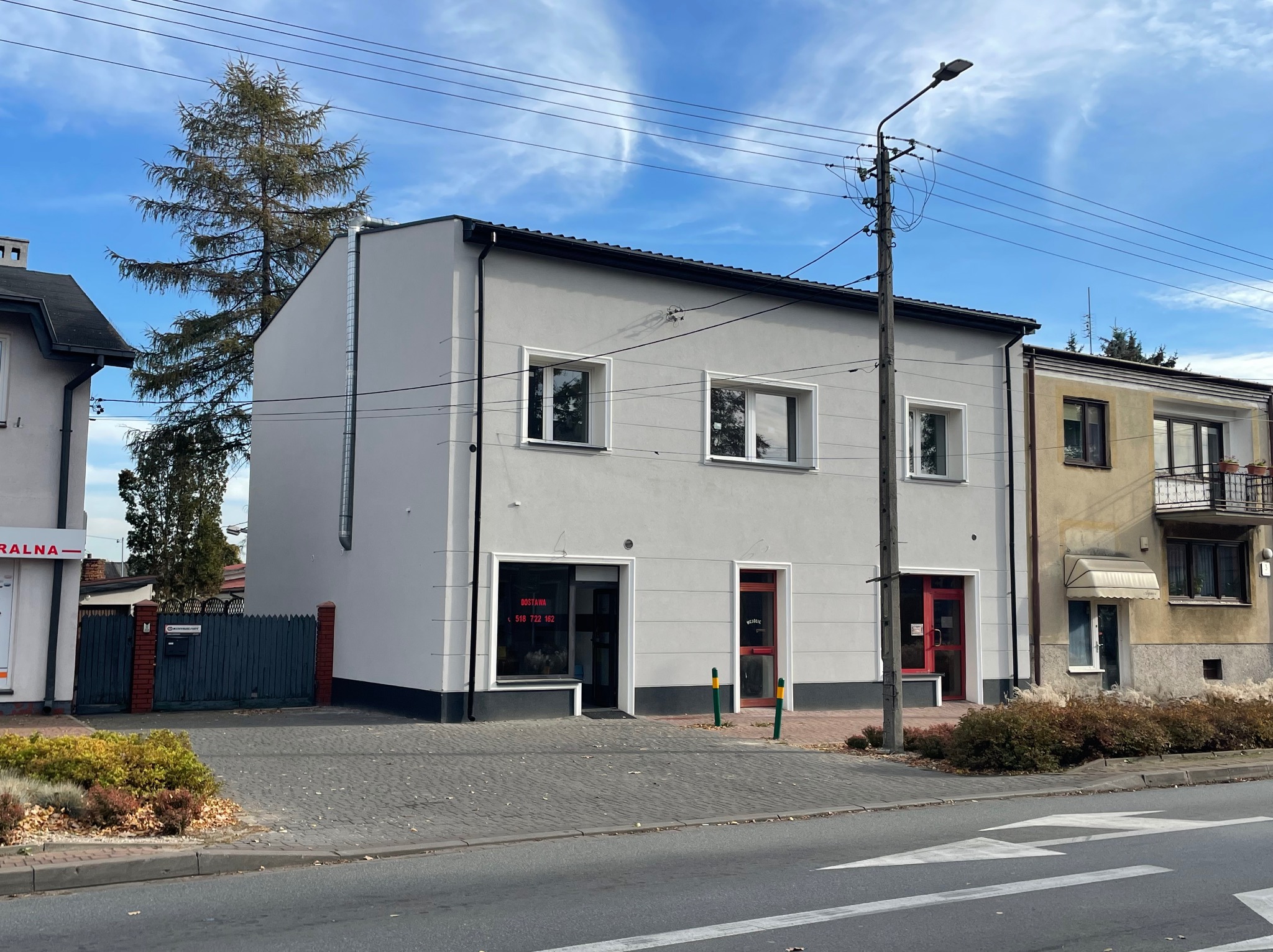 Dwukondygnacyjny budynek po odnowieniu elewacji, pomalowany na szaro z białymi obramowaniami okien i czerwonymi drzwiami wejściowymi, widoczny z poziomu ulicy w słoneczny dzień.