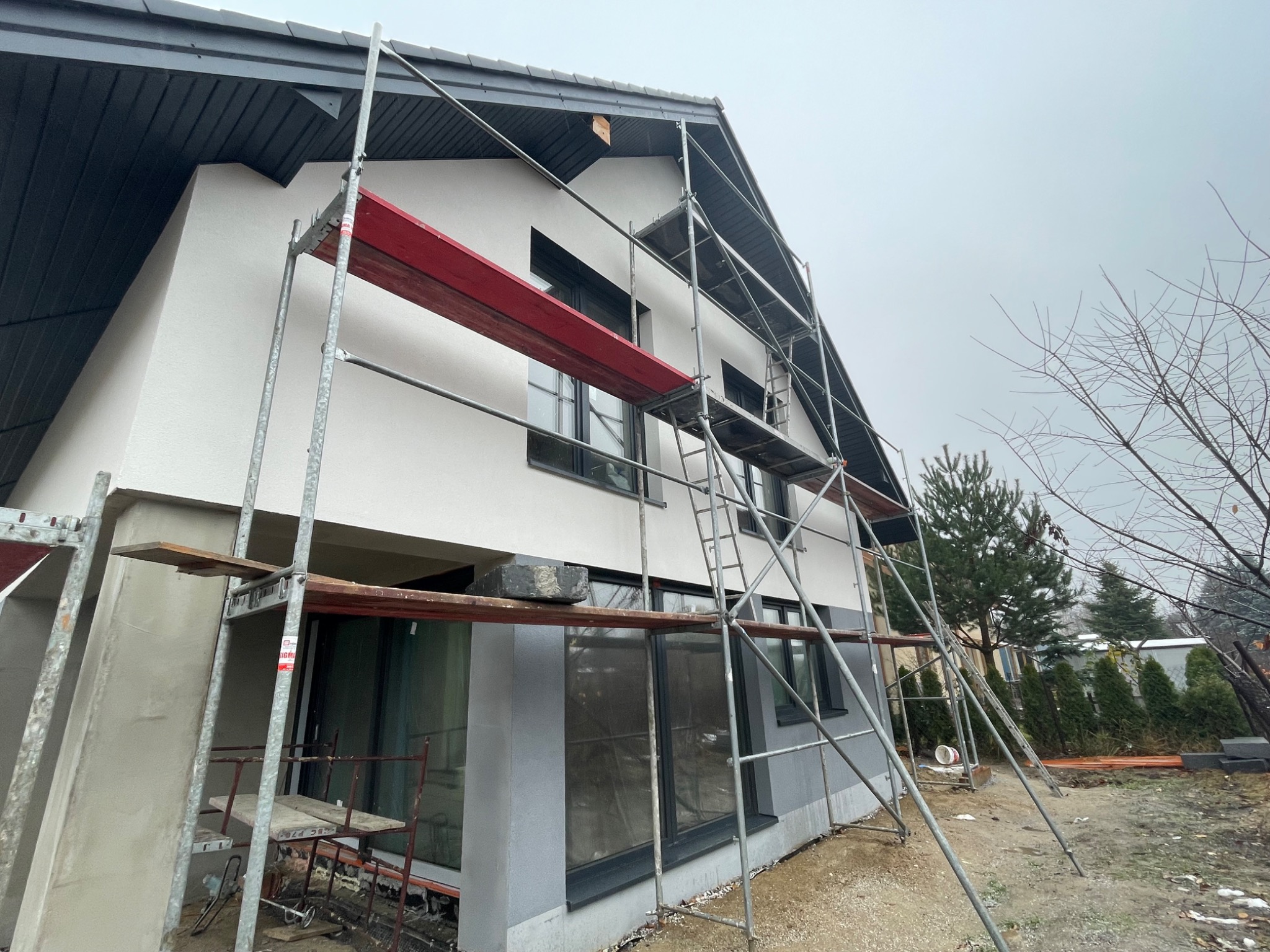 Dom w trakcie prac elewacyjnych, widoczne rusztowania, częściowo otynkowana i pomalowana fasada, szara stolarka okienna, pochmurne niebo.