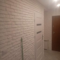 R-F Remonty - Ściana z białej cegły dekoracyjnej, białe drzwi wewnętrzne z poziomymi przeszkleniami, brązowe drzwi wejściowe w tle, widoczny fragment grzejnika.