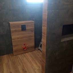 R-F Remonty - Ciemnoszara łazienka z drewnopodobną zabudową stelaża WC, widoczne rury odpływowe i podłączenie do spłuczki, drewnopodobna podłoga, drabina w tle.