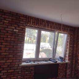 R-F Remonty - Pokój po ułożeniu cegieł dekoracyjnych na ścianach wokół okna, narzędzia i wiadro kleju na parapecie, widok na zewnątrz.