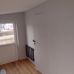 R-F Remonty - Pomalowane na szaro wnętrze pokoju z drewnianą podłogą, białymi drzwiami balkonowymi i ukrytymi drzwiami wewnętrznymi z czarną klamką, obok których zamontowano składany grzejnik.
