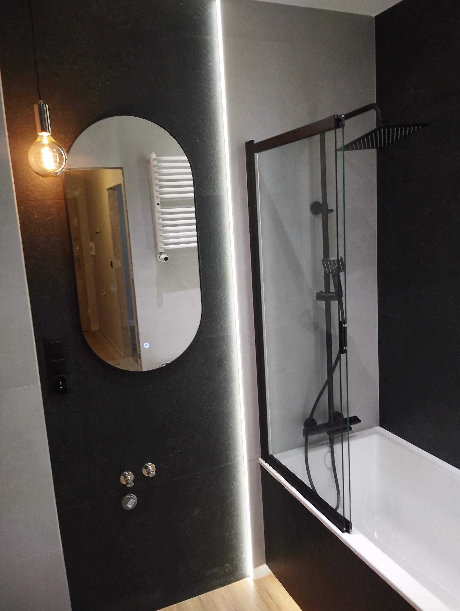 Nowoczesna łazienka z czarną armaturą: owalne lustro z podświetleniem LED, kabina prysznicowa nad wanną i ciemne płytki. Widoczne detale wykończenia.