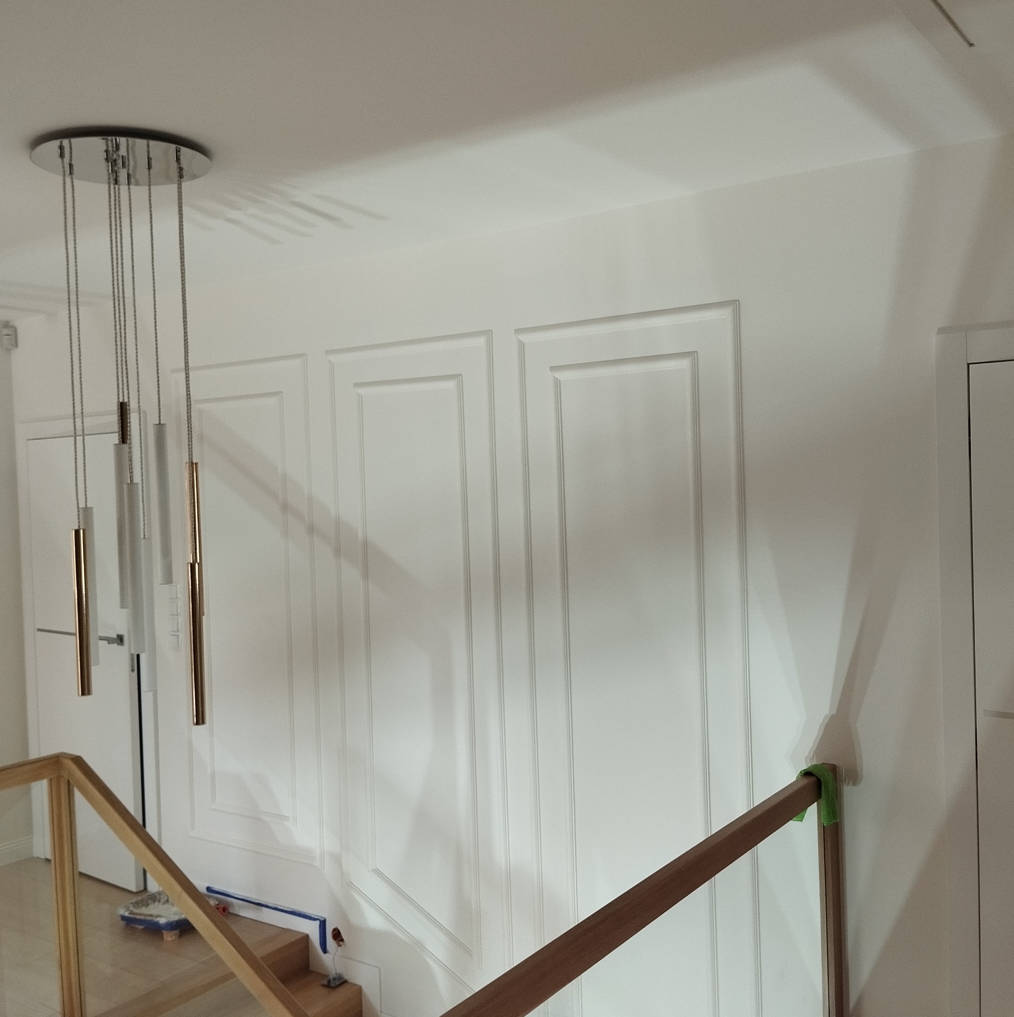 Białe ściany z dekoracyjnymi panelami i nowoczesną lampą wiszącą. Widoczna balustrada schodów i drzwi. Detal wykończenia wnętrza w jasnym, minimalistycznym stylu.