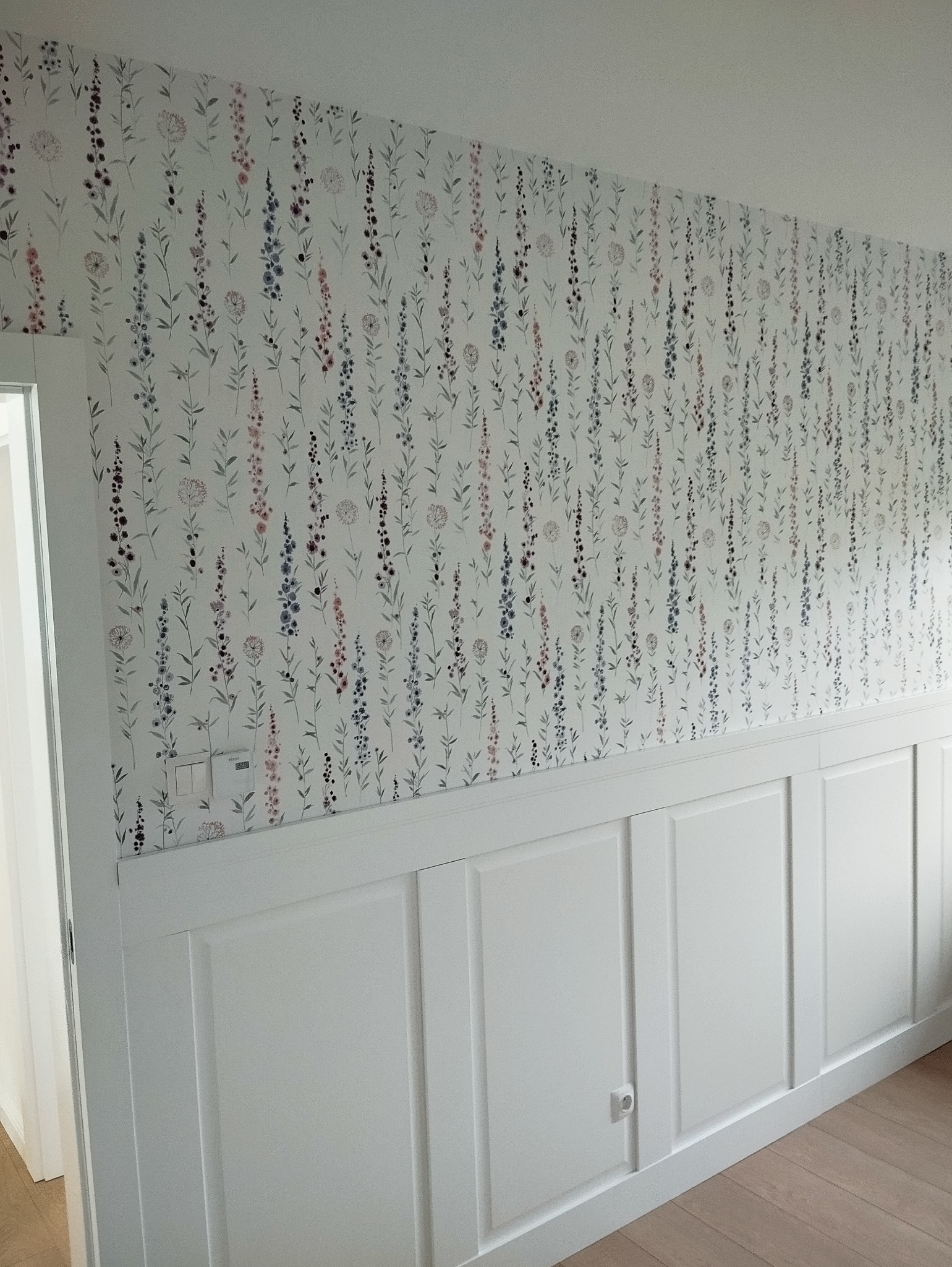 Ściana z białą boazerią i tapetą w drobne, pastelowe kwiaty. Widoczny fragment podłogi z jasnego drewna i drzwi po lewej stronie kadru. Subtelny wzór tapety.