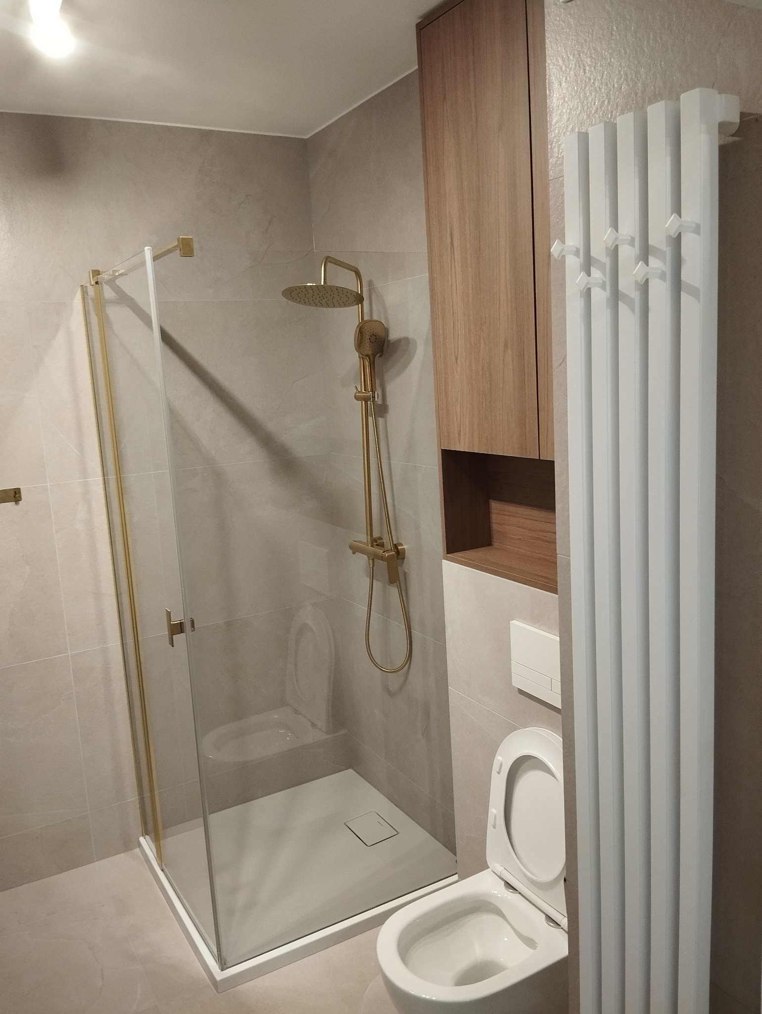 Wykończona łazienka z kabiną prysznicową ze złotymi elementami, grzejnikiem drabinkowym i toaletą. Jasne płytki na ścianach i podłodze. Drewniana szafka nad toaletą.