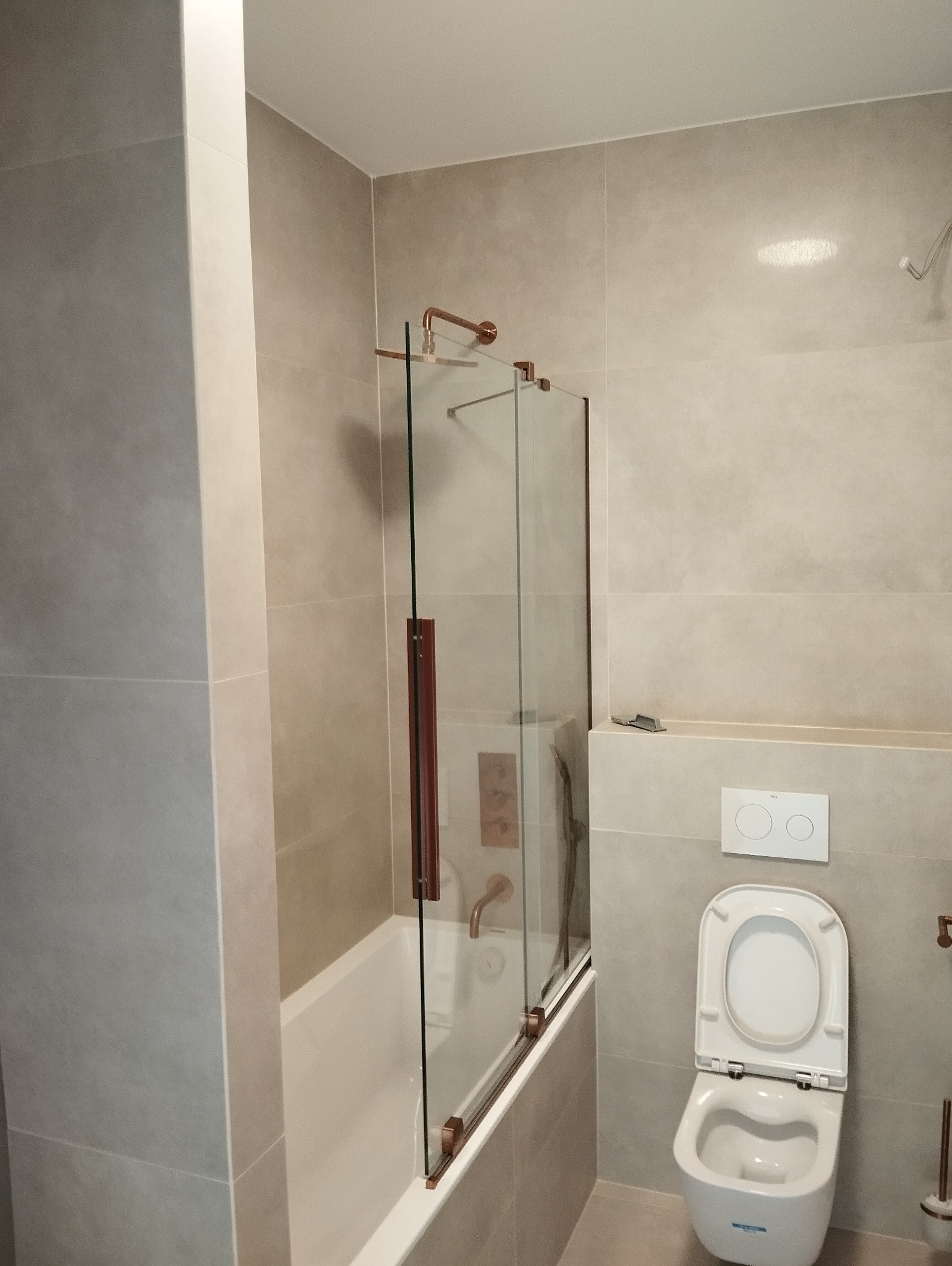 Nowoczesna łazienka w beżowych płytkach z wanną i szklanym parawanem z miedzianymi detalami oraz wiszącą toaletą. Minimalistyczny design.