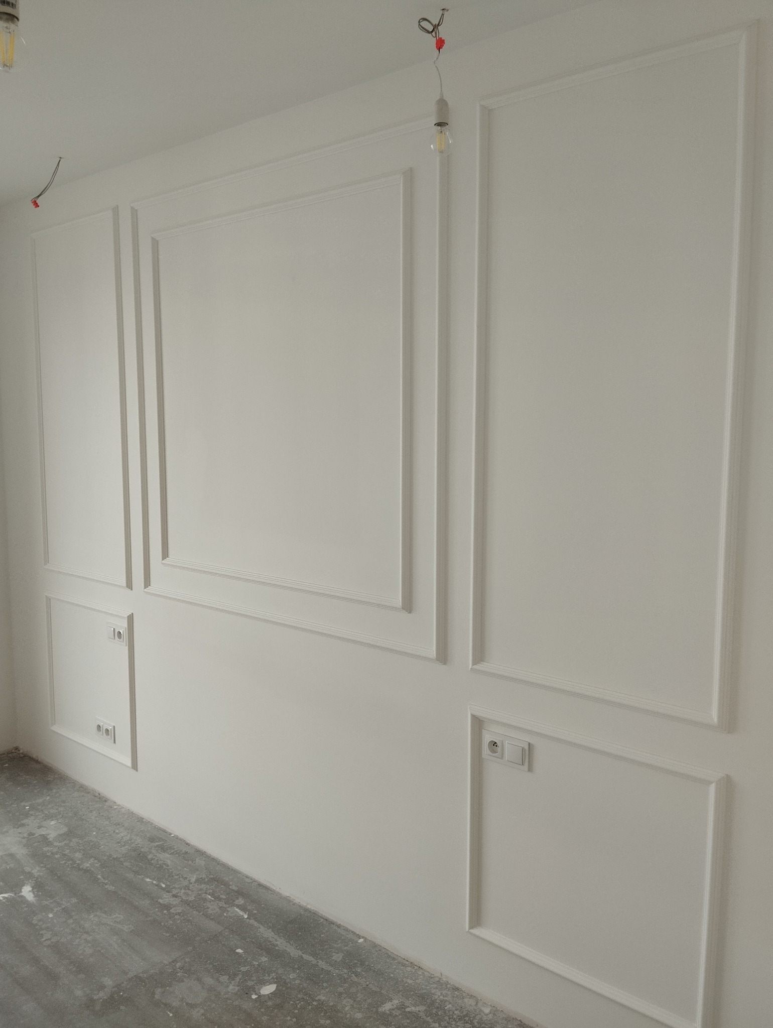 Biała ściana z dekoracyjnymi panelami i gniazdkami elektrycznymi. Widoczne niedokończone podłoże podłogowe i wisząca żarówka.