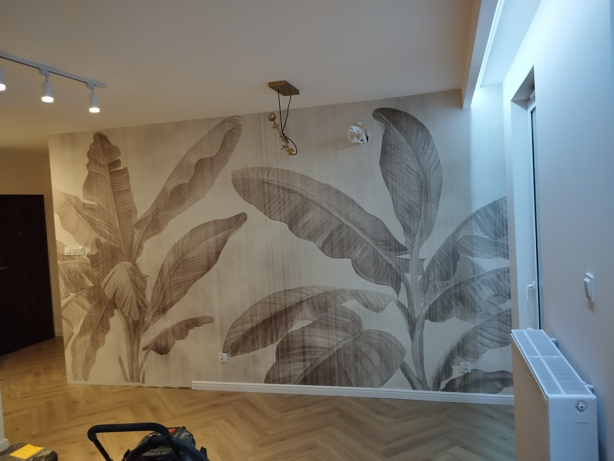 Wnętrze po wykończeniu: ściana z motywem liści bananowca, parkiet w jodełkę, białe listwy przypodłogowe i grzejnik. Widoczne lampy sufitowe i fragment odkurzacza.