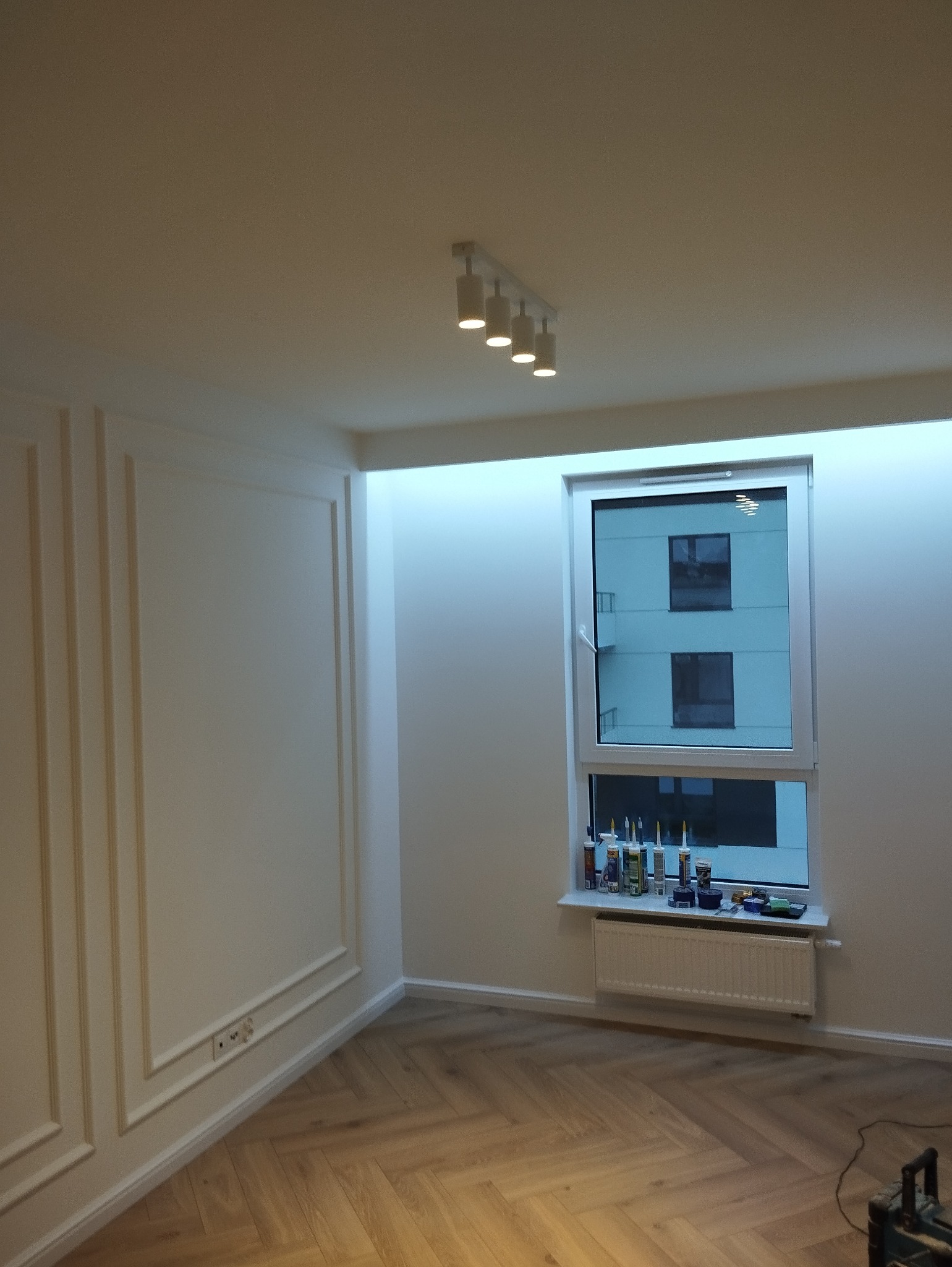 Jasne wnętrze po remoncie: parkiet w jodełkę, białe ściany z dekoracyjnymi listwami, okno z widokiem na budynek. Nowoczesne oświetlenie sufitowe.