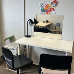 Gabinet terapeutyczny z białym biurkiem, czarną lampką, lustrem i naklejką z postaciami z bajki Psi Patrol na ścianie, widoczne dwa czarne krzesła i roślina doniczkowa.