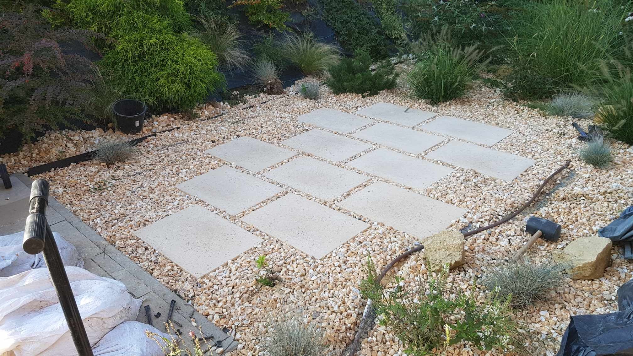 Układanie ścieżki z jasnych, prostokątnych płyt na podłożu z drobnych, beżowych kamieni. W tle widoczne rośliny ogrodowe i czarny wiadro.