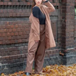 Kobieta w brązowym płaszczu i spodniach pozuje na tle ceglanej ściany, otoczona opadłymi liśćmi.