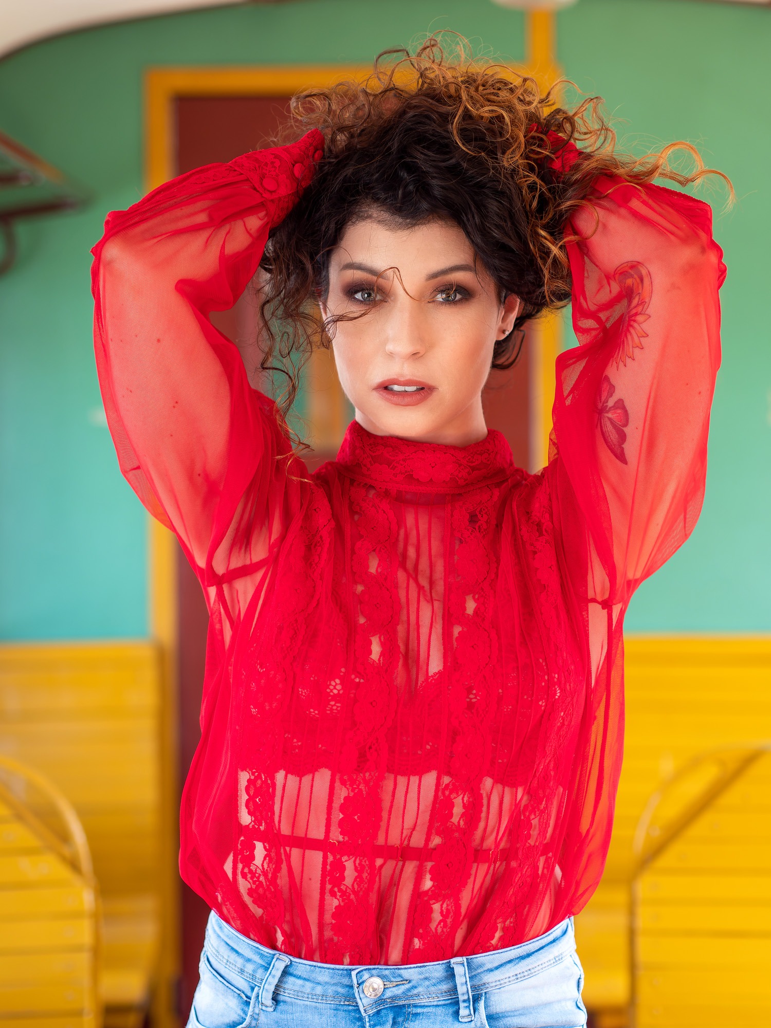 Portret kobiety w czerwonej, koronkowej bluzce, z tatuażem motyla na ramieniu, dłońmi uniesionymi do góry, z kręconymi włosami na tle kolorowej ściany.