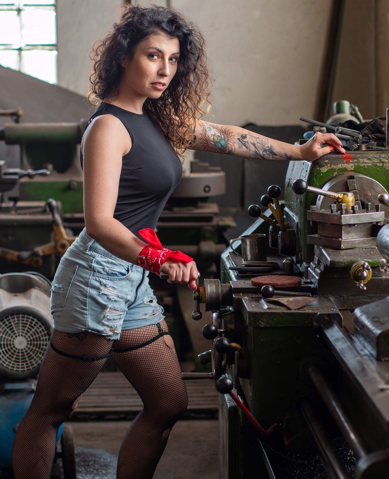 Kobieta z tatuażami w dżinsowych szortach i kabaretkach, z czerwoną bandaną na nadgarstku, obsługuje starą tokarkę w warsztacie.