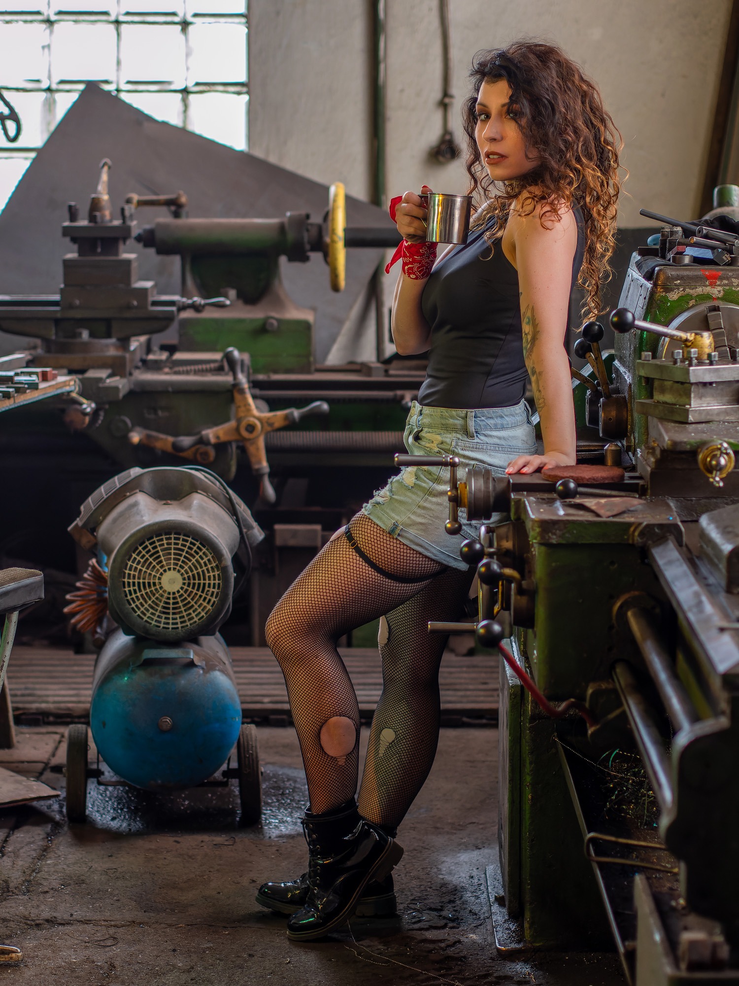 Kobieta z kręconymi włosami, w czarnym topie i dżinsowych szortach, oparta o starą tokarkę w warsztacie, trzyma metalowy kubek.