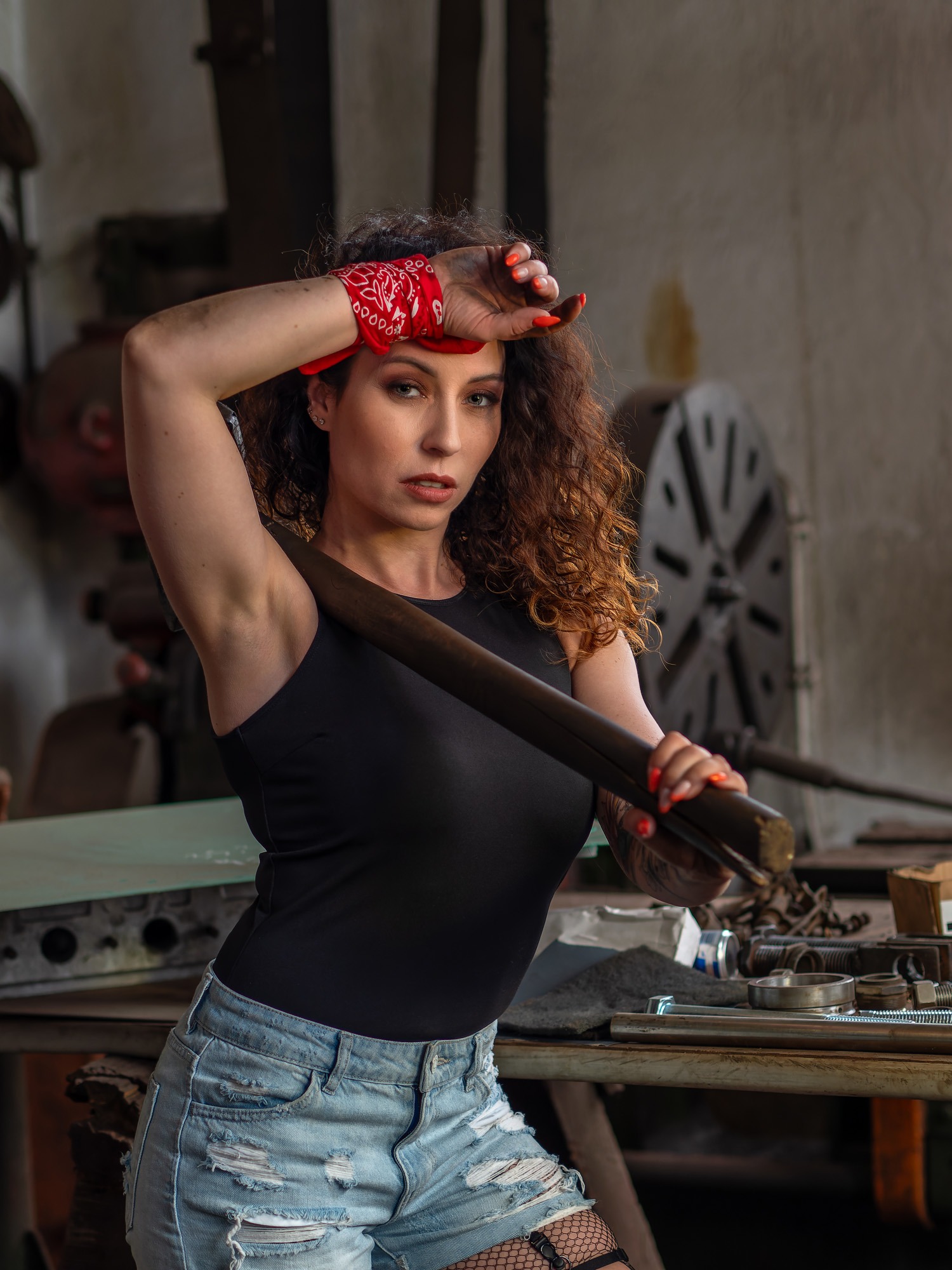 Kobieta z czerwoną bandaną we włosach, w czarnym topie i jeansowych szortach, opiera się o metalowy pręt na tle warsztatu z narzędziami i maszynerią.