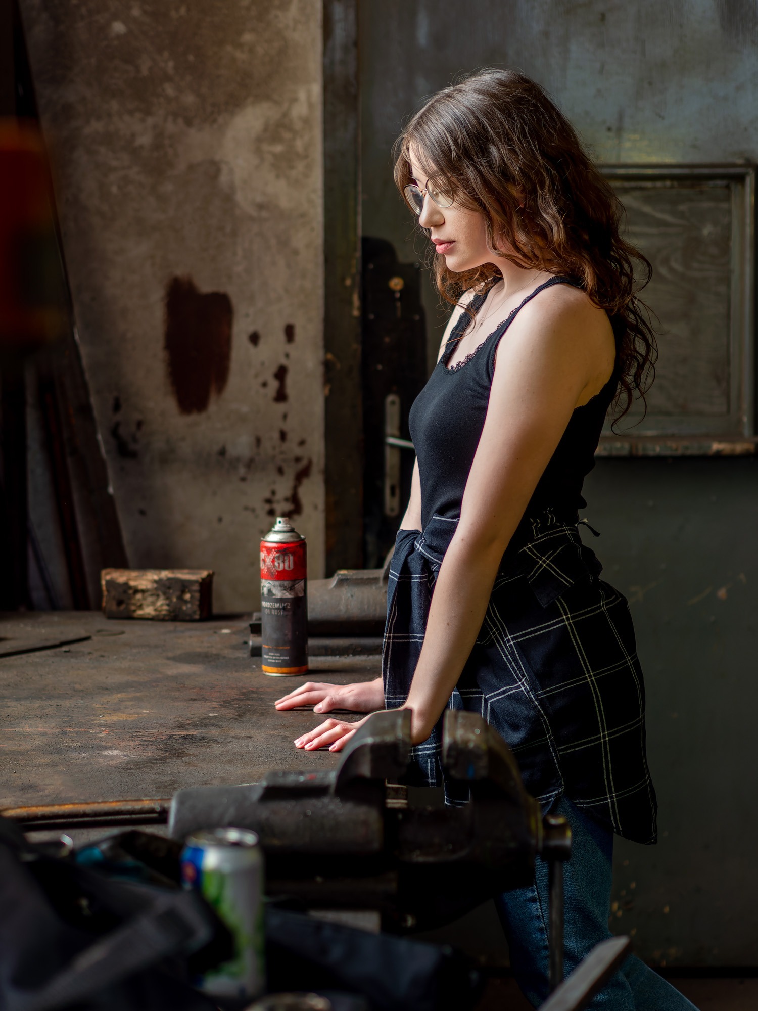 Kobieta w okularach, w warsztacie, opierająca się o stół z imadłem i puszką smaru, w tle okno i ściana z zaciekami.