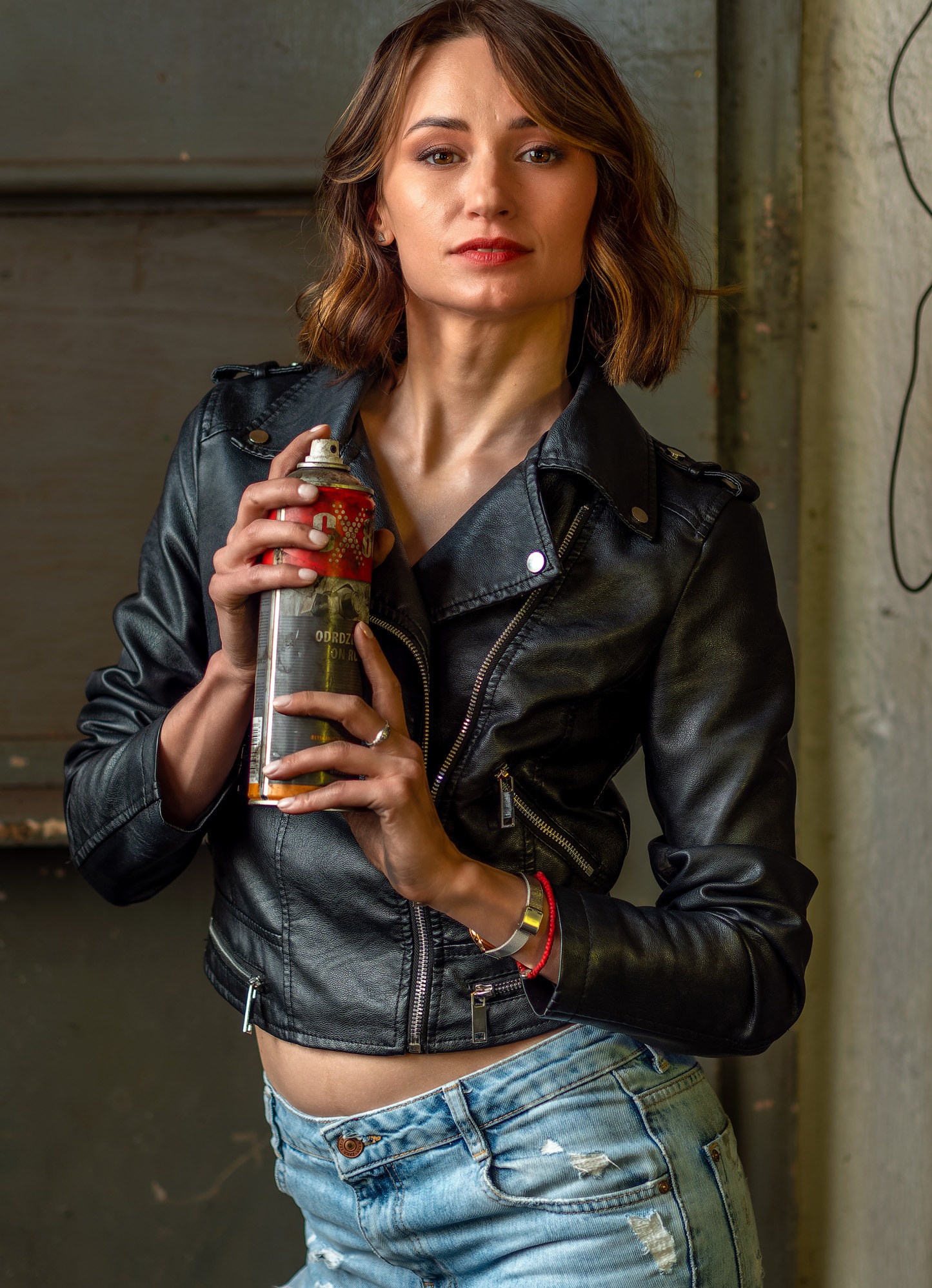 Kobieta w skórzanej kurtce trzyma puszkę ze sprayem na tle szarej ściany.