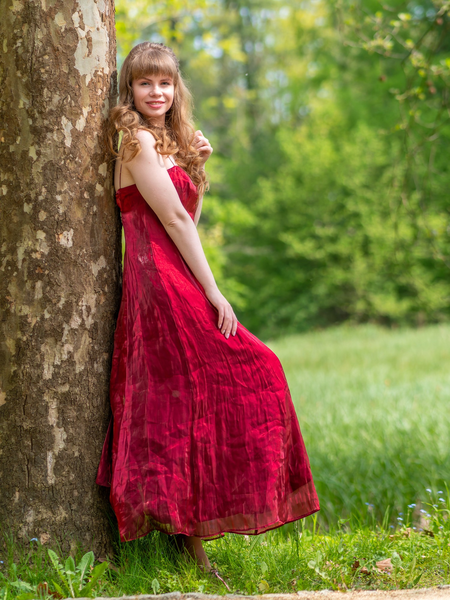 Portret młodej kobiety w długiej czerwonej sukni, opierającej się o drzewo w zielonym parku.