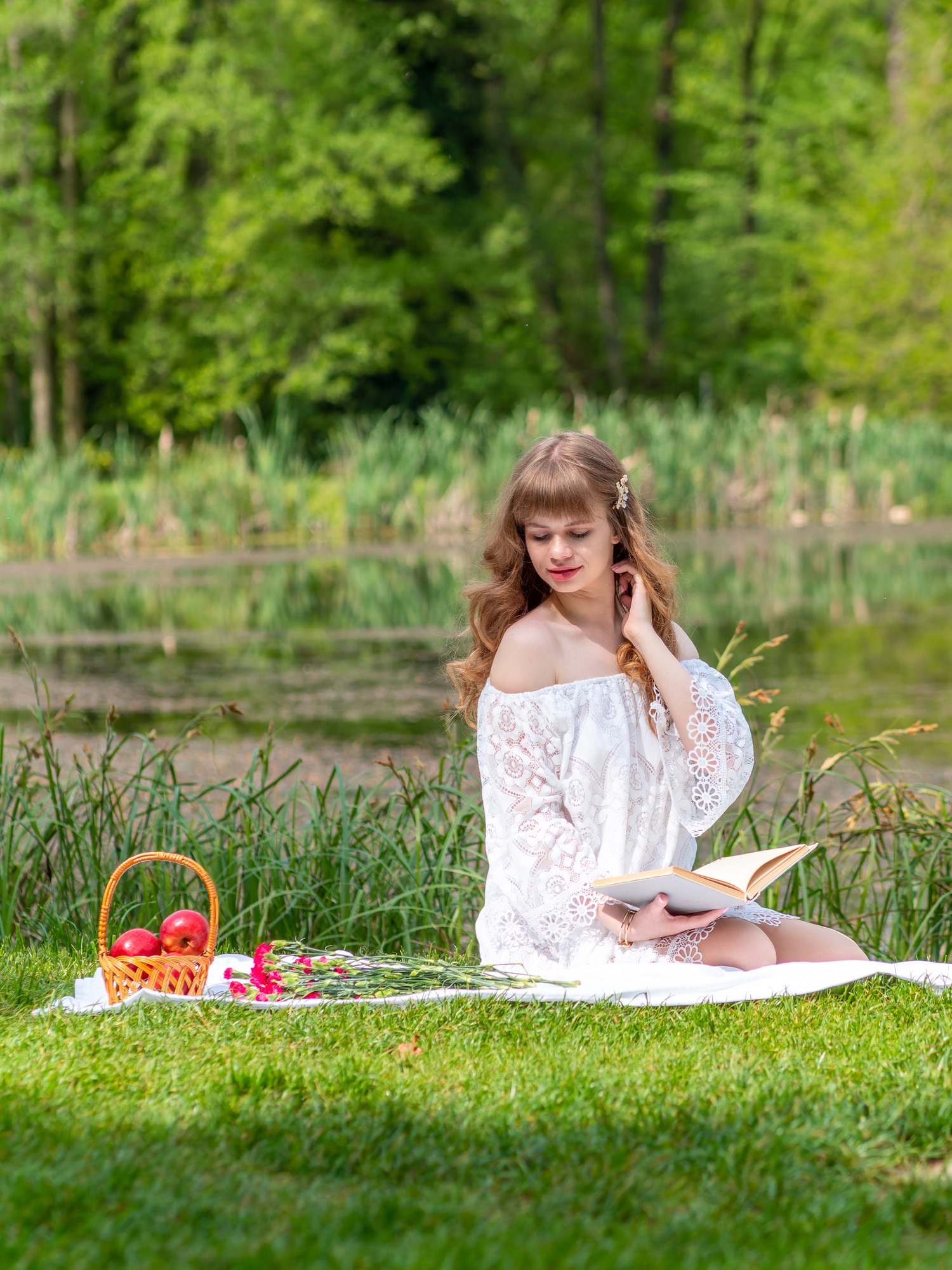 Młoda kobieta w białej koronkowej sukience czyta książkę na pikniku nad brzegiem jeziora, obok koszyk z jabłkami i goździki.