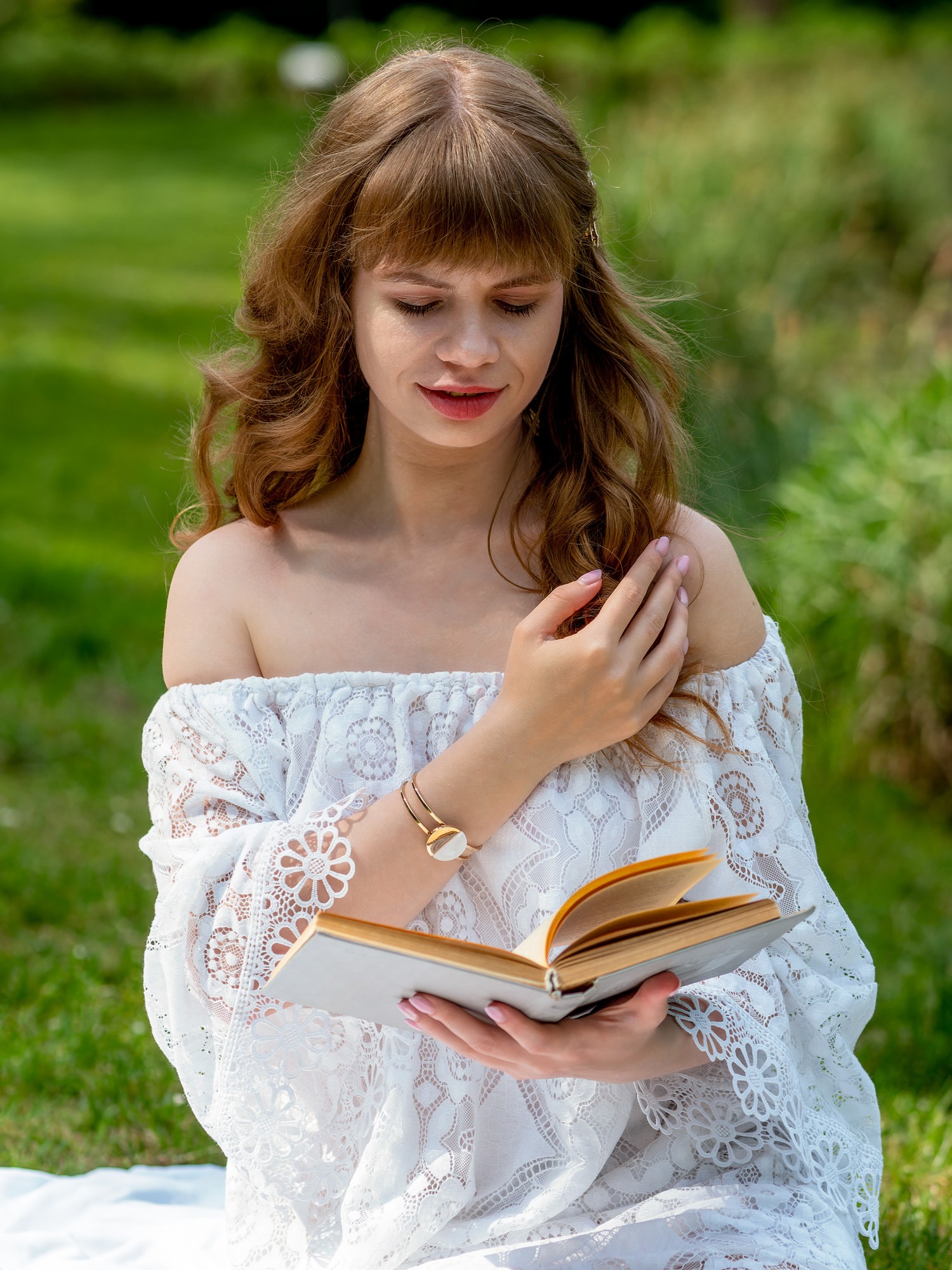 Portret kobiety z rudymi włosami w białej koronkowej sukience, czytającej książkę na trawie w parku.