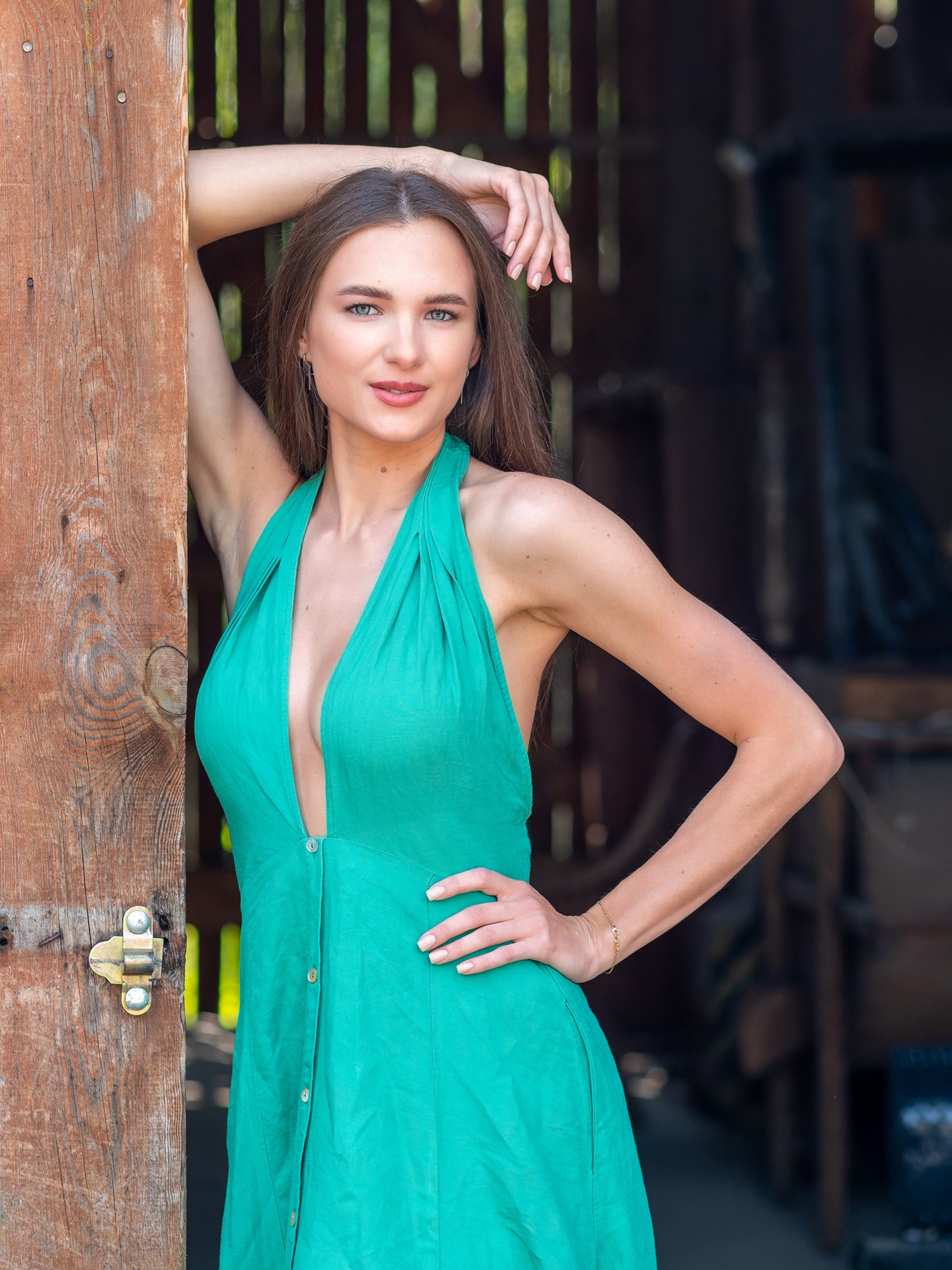 Portret kobiety w zielonej sukience pozującej przy drewnianych drzwiach stodoły, z widocznym złotym łańcuszkiem na nadgarstku i delikatnym makijażem.