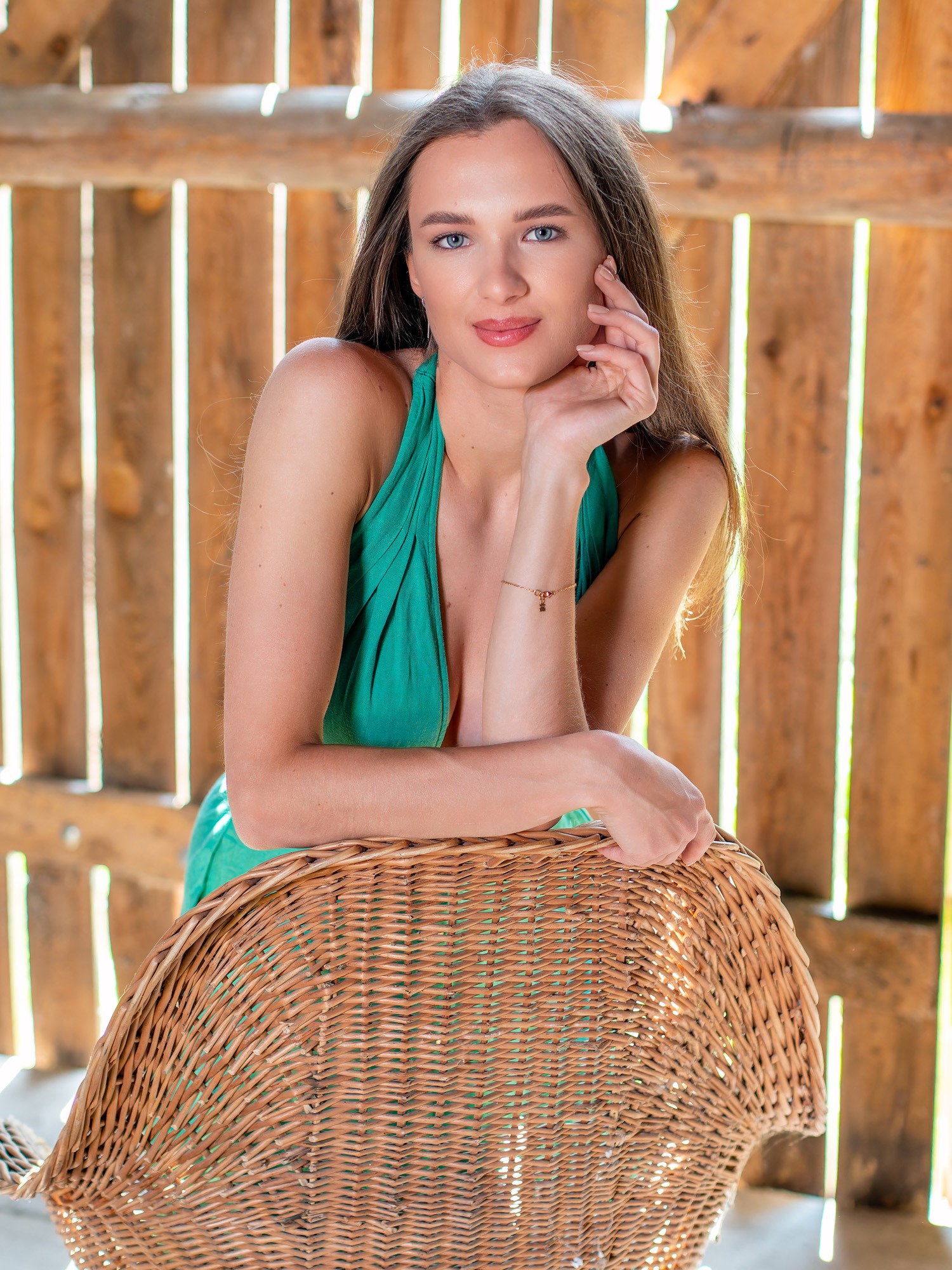 Portret kobiety w zielonej sukience, opierającej się o wiklinowy fotel na tle drewnianej konstrukcji, z delikatną bransoletką na nadgarstku.