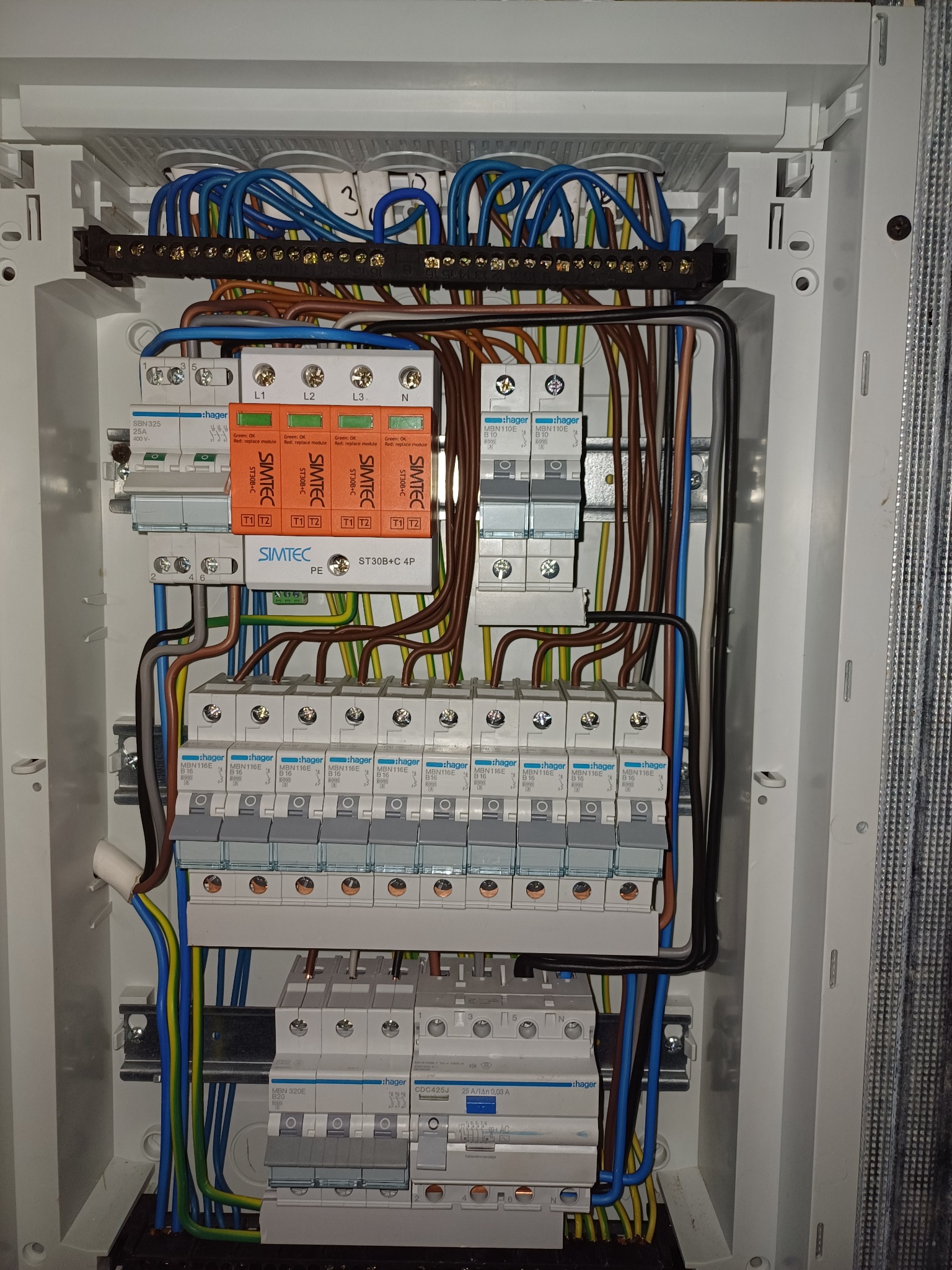 Otwarta skrzynka elektryczna z widocznymi bezpiecznikami, przewodami różnej grubości i kolorów, oraz ochronnikami przepięć firmy SIMTEC, zamontowanymi na szynie DIN.