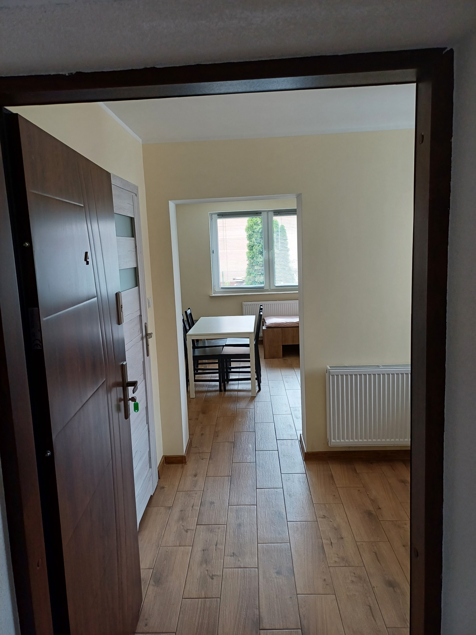 Widok z korytarza na odnowione wnętrze mieszkania z nową podłogą imitującą drewno, grzejnikiem, drzwiami wejściowymi i pokojem z białym stołem oraz oknem z widokiem na zieleń.