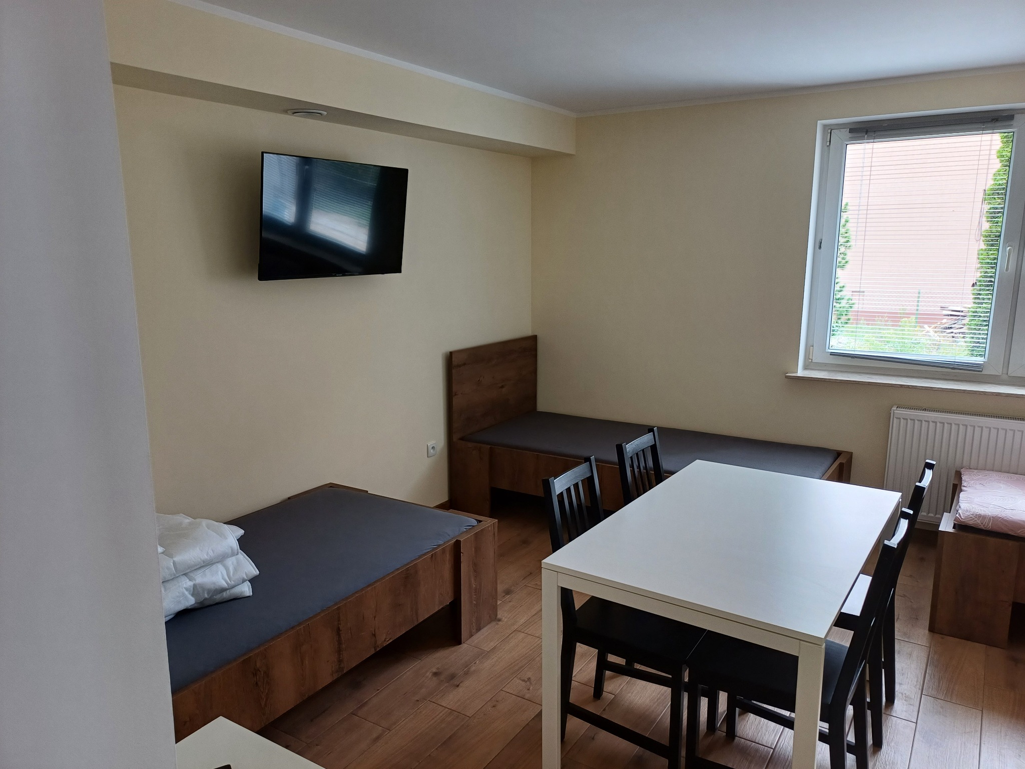 Jasne wnętrze pokoju z dwoma łóżkami z drewnianymi ramami i szarą pościelą, białym stołem z czarnymi krzesłami oraz telewizorem LCD zawieszonym na ścianie.