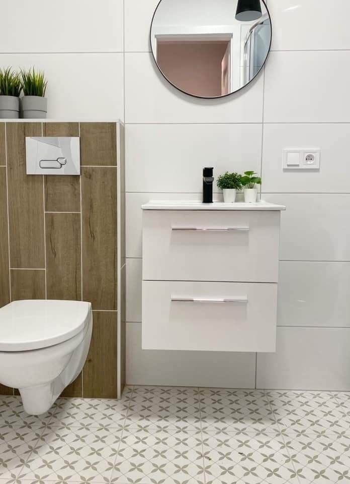 Nowoczesna łazienka po remoncie: białe płytki, wisząca szafka z umywalką, czarna bateria, okrągłe lustro i wzorzysta podłoga.