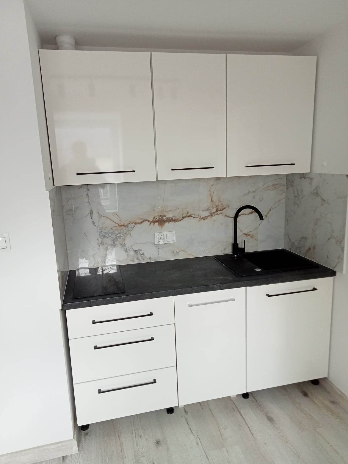 Nowoczesna, biała zabudowa kuchenna z czarnym blatem i armaturą, marmurowy backsplash, widoczne gniazdko elektryczne.