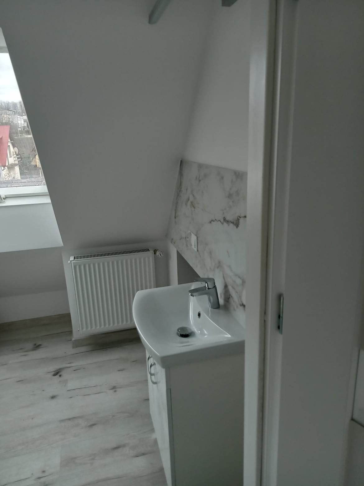 Biała umywalka z chromowaną baterią w łazience na poddaszu, z marmurową okładziną ścienną i grzejnikiem żeberkowym w tle, widok zza otwartych drzwi.