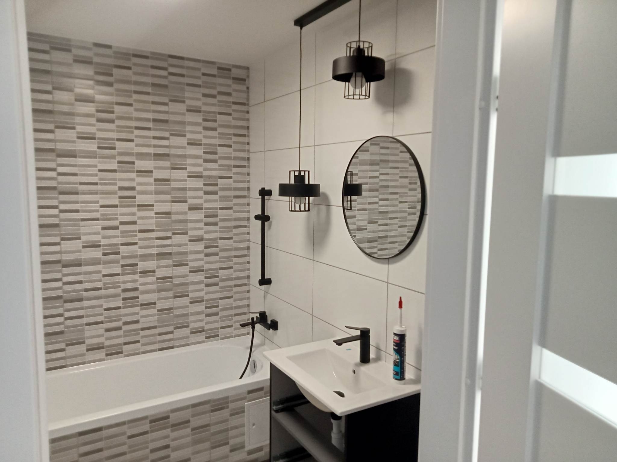 Wykończona łazienka z wanną obłożoną mozaiką, czarną armaturą, okrągłym lustrem i nowoczesnymi lampami wiszącymi, widoczna tuba silikonu sanitarnego na blacie umywalki.