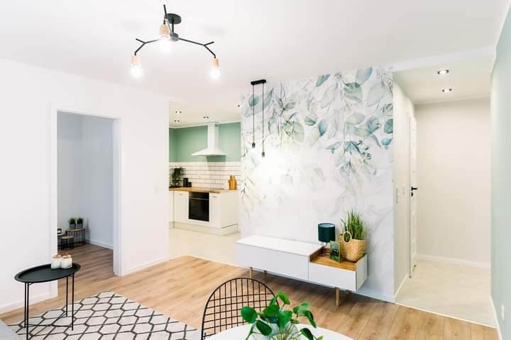 Jasne wnętrze mieszkania po remoncie: parkiet, białe ściany, fototapeta z motywem liści, nowoczesna lampa sufitowa i wisząca, widok na kuchnię z zielonymi akcentami.