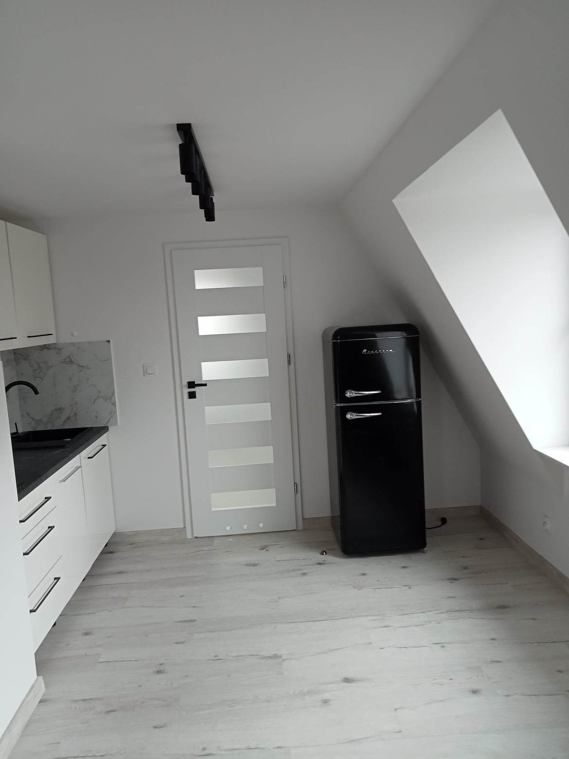 Nowoczesna kuchnia w bieli i czerni z oświetleniem szynowym, czarną lodówką w stylu retro i białymi frontami szafek z czarnymi uchwytami.