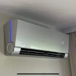 Nowoczesny, srebrny klimatyzator ścienny z niebieskim podświetleniem LED, zamontowany w jasnym pomieszczeniu z kremowymi ścianami i zasłonami.