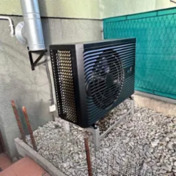 Czarna pompa ciepła powietrze-woda, zamontowana na zewnątrz budynku na metalowej konstrukcji, otoczona kamieniami dekoracyjnymi i siatką ogrodzeniową, z widocznym kominem wentylacyjnym i fragmentem...