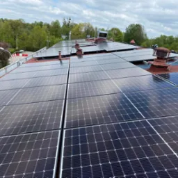 Rozległa instalacja paneli słonecznych na dachu budynku z czerwonym pokryciem, widoczne kominy wentylacyjne i anteny, w tle zieleń drzew i pochmurne niebo.