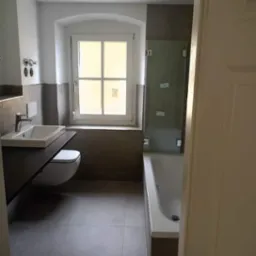 Nowoczesna łazienka z szarymi płytkami, wiszącą toaletą i umywalką na blacie, kabina prysznicowa nad wanną, okno z łukiem.